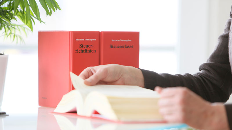 Motiv Steuerberatung, Blick auf den Schreibtisch, Bücher Steuerrichtlinien, Steuererlasse, unscharfer Vordergrund, Person liest im BUch